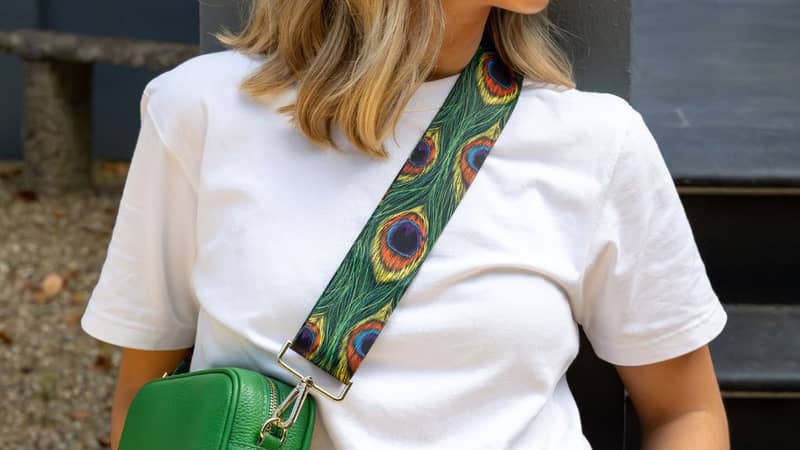 Blonde model wearing sunglasses white shirt black trousers gold jewellery with a peacock bag strap a green crossbody italian leather bag