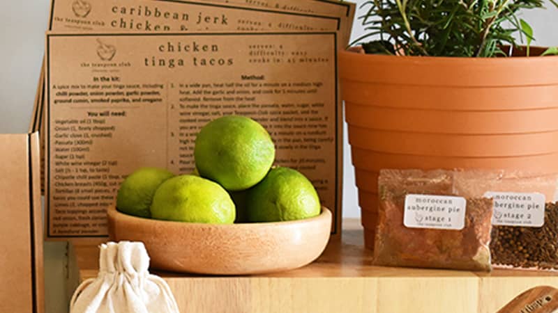 Fresh, potted thyme and recipe cards sit on a shelf, with spice packets, a bowl filled with turmeric, and limes in a dish in the foreground