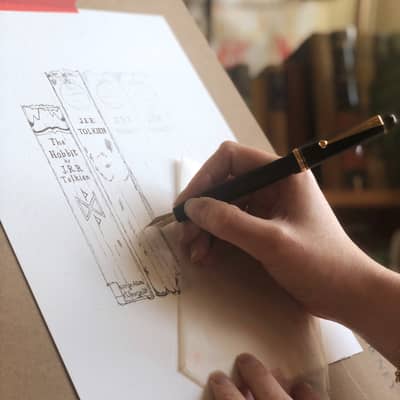 Georgina's hand holding a black fountain pen sketches a book spine onto a piece of paper mounted on an easel, a shelf of books can be seen in the background