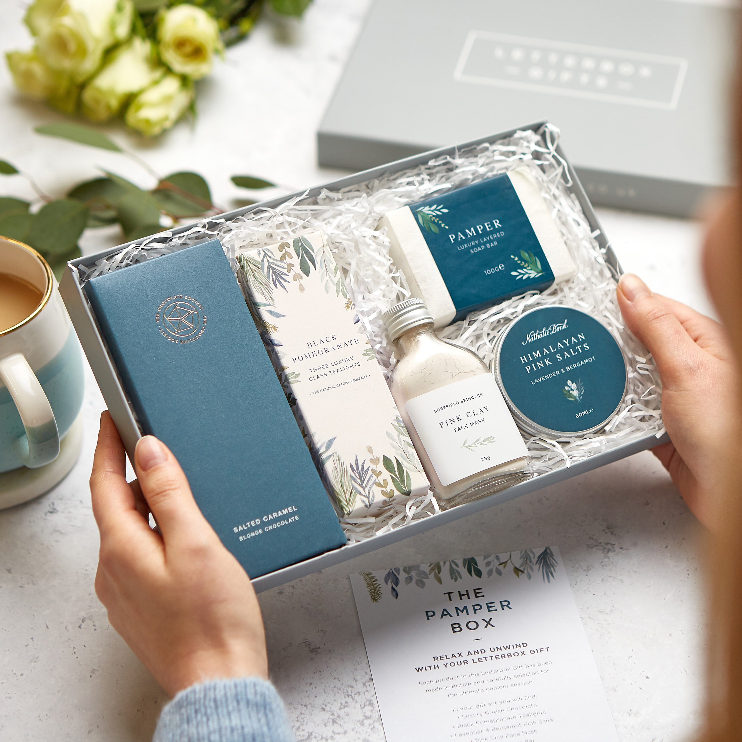 A pampering gift set in a grey presentation gift box containing salted caramel chocolate, black pomegranate tealights, pink clay face mask, Himalayan bath salts