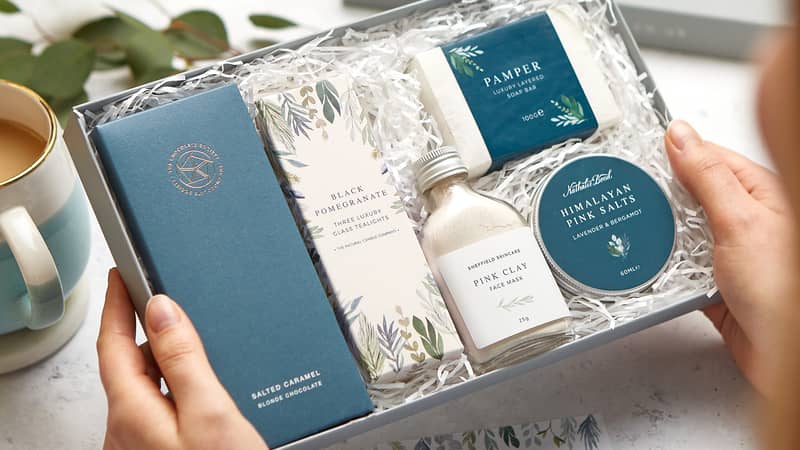 A pampering gift set in a grey presentation gift box containing salted caramel chocolate, black pomegranate tealights, pink clay face mask, Himalayan bath salts