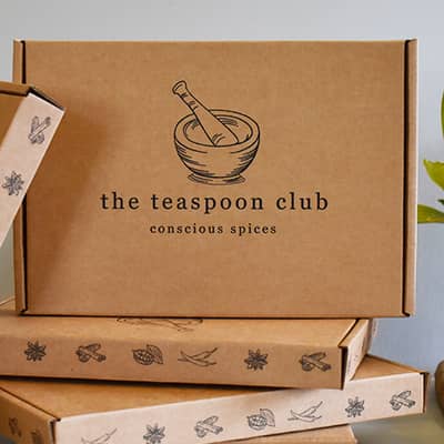 A printed box sits atop 3 stacked boxes. The stacked boxes show illustrated spices along the trim, with a pessle and mortar Teaspoon Club logo on the front