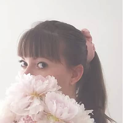 Headshot of Jo holding pink peonies over her lower face with a pink silk scrunchie in her ponytail