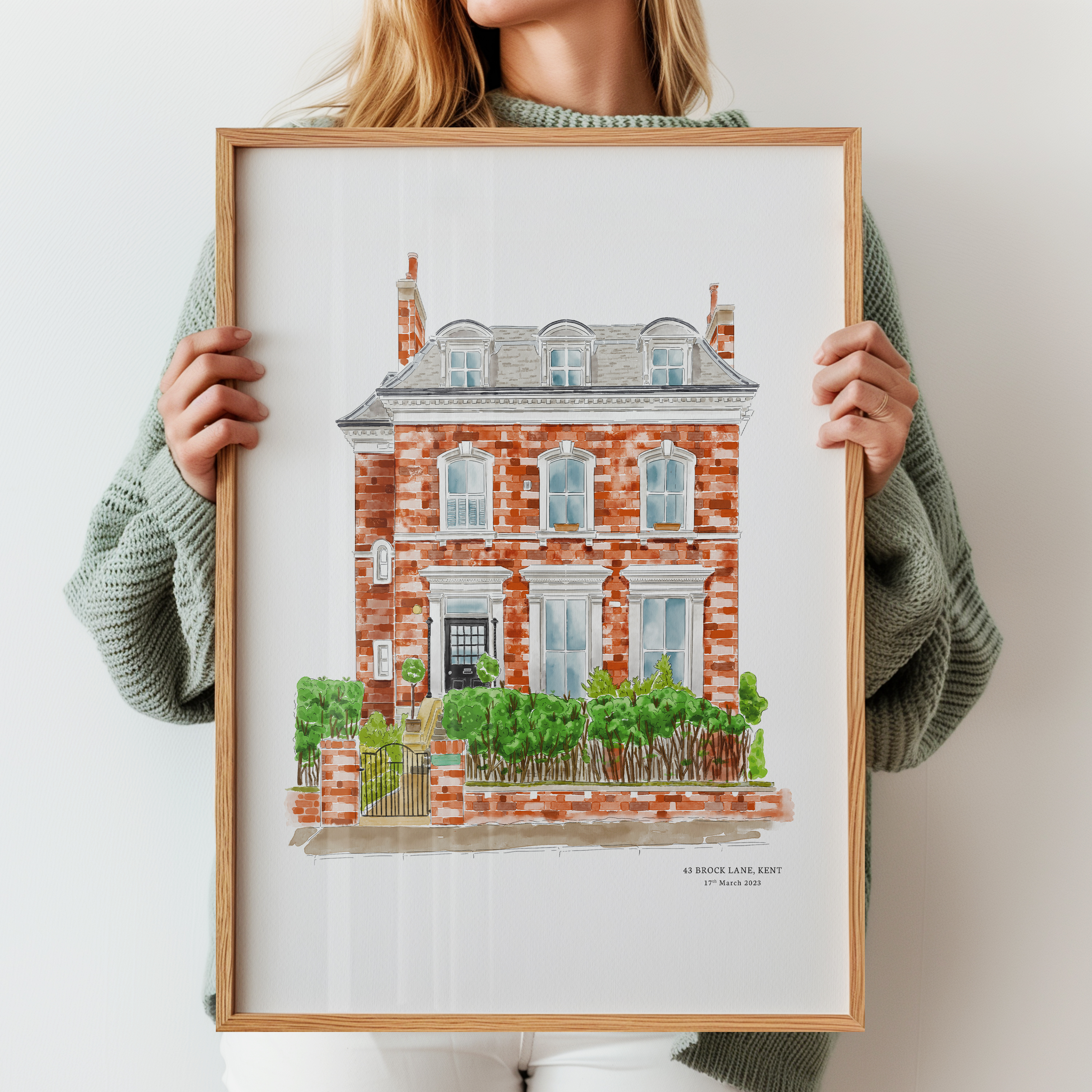 A beautifully detailed watercolor-style illustration of a red-brick house, framed and held by a person in a cozy sweater.