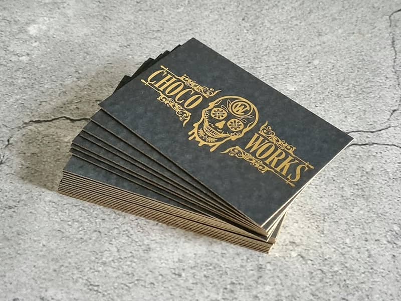 A stack of black business cards on a stone-grey background. The business cards include gold-foil edges and gold-foil logo
