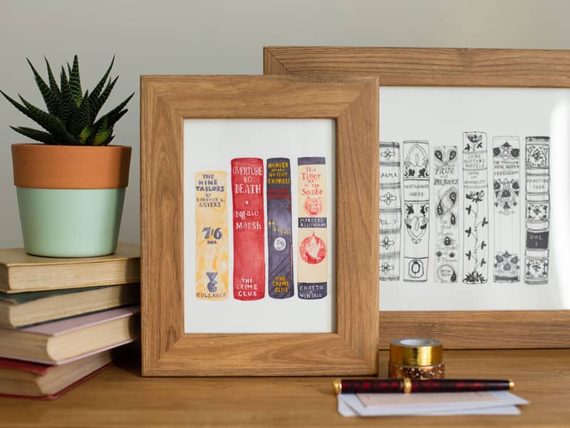 Two book spine artworks, in oak frames, displayed next to a stack of books.