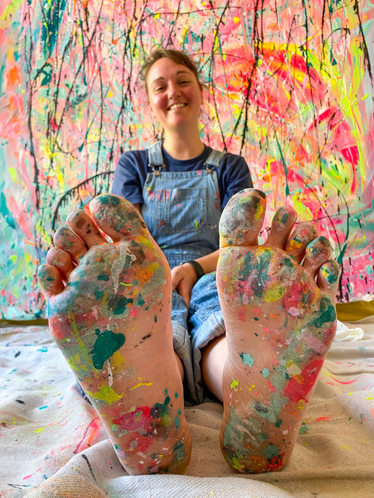 Michelle, artist and creator behind Eat Pray Pedal sitting in front of a large canvas with paint splattered feet.