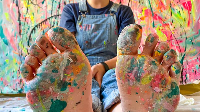 Michelle, artist and creator behind Eat Pray Pedal sitting in front of a large canvas with paint splattered feet.