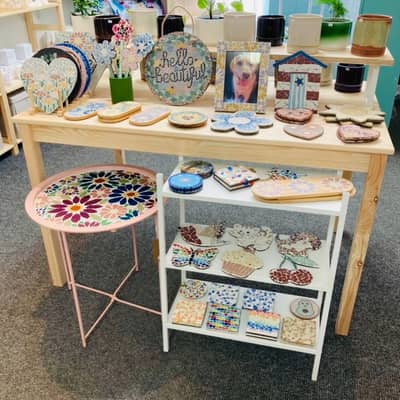 A picture of Hayleys Mosaics items in a shop. Colourful mosaic items including wreaths, photo frames and coasters are stacked on shelves