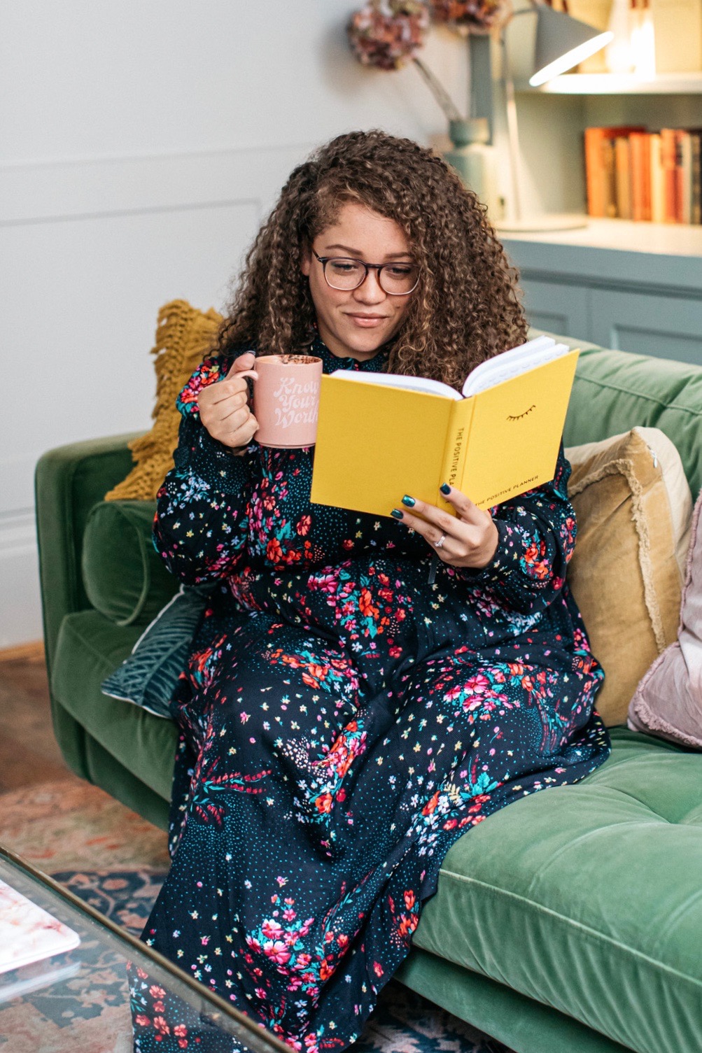 A lady sat on a sofa with a cup of coffee reading a bright yellow The Positive Planner journal 