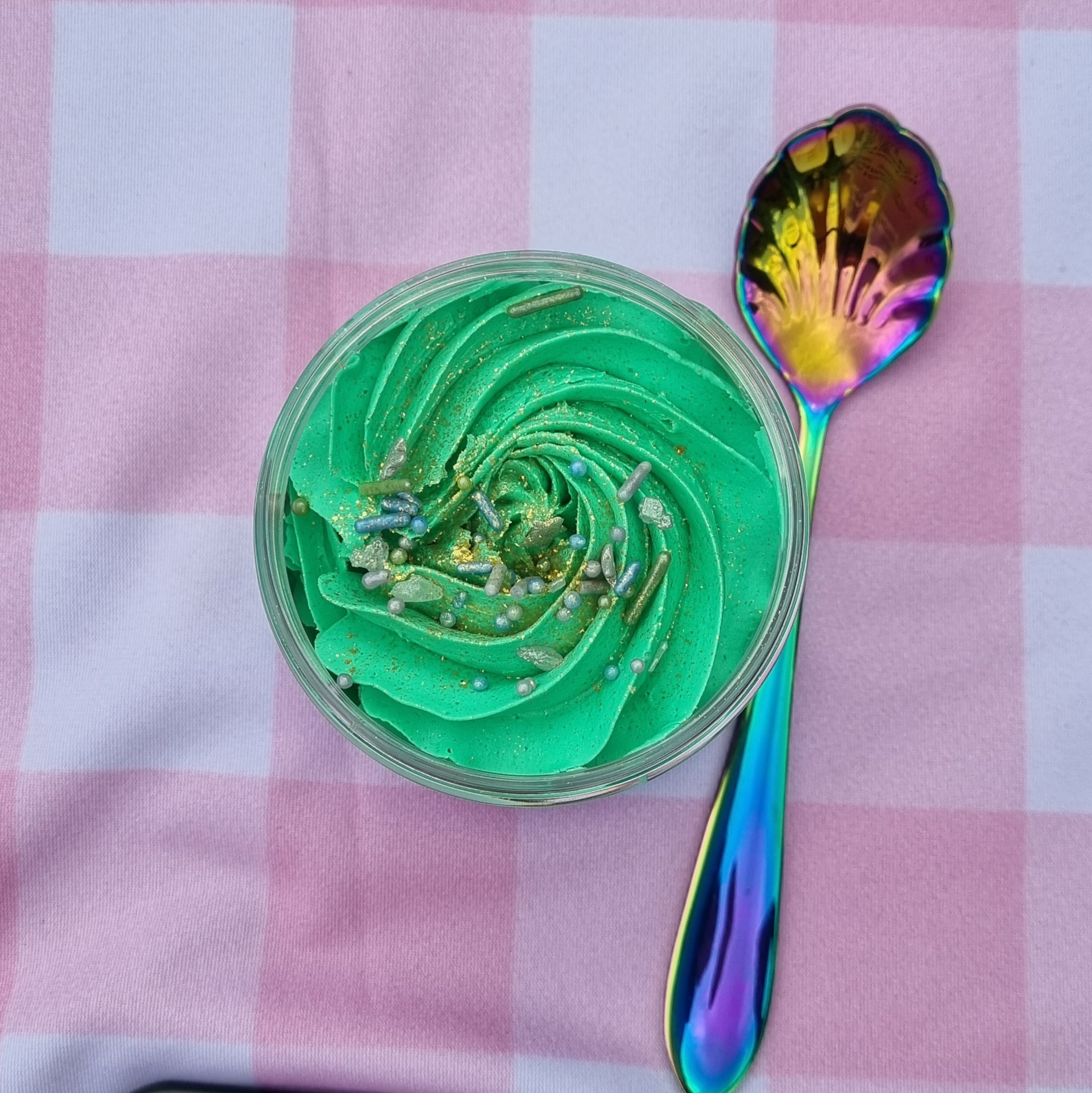 A pastel pink check background with a pot of green body wash that has been whipped with a spoon next to it