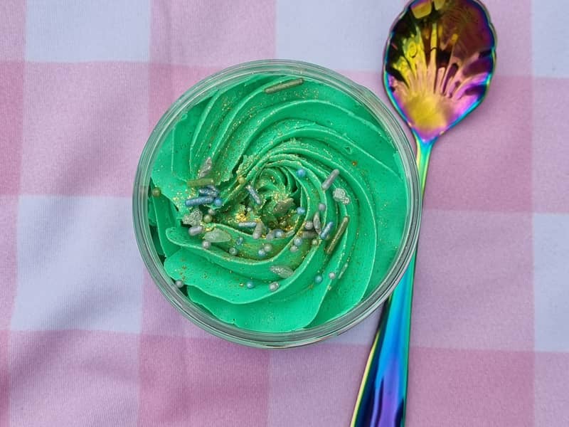 A pastel pink check background with a pot of green body wash that has been whipped with a spoon next to it