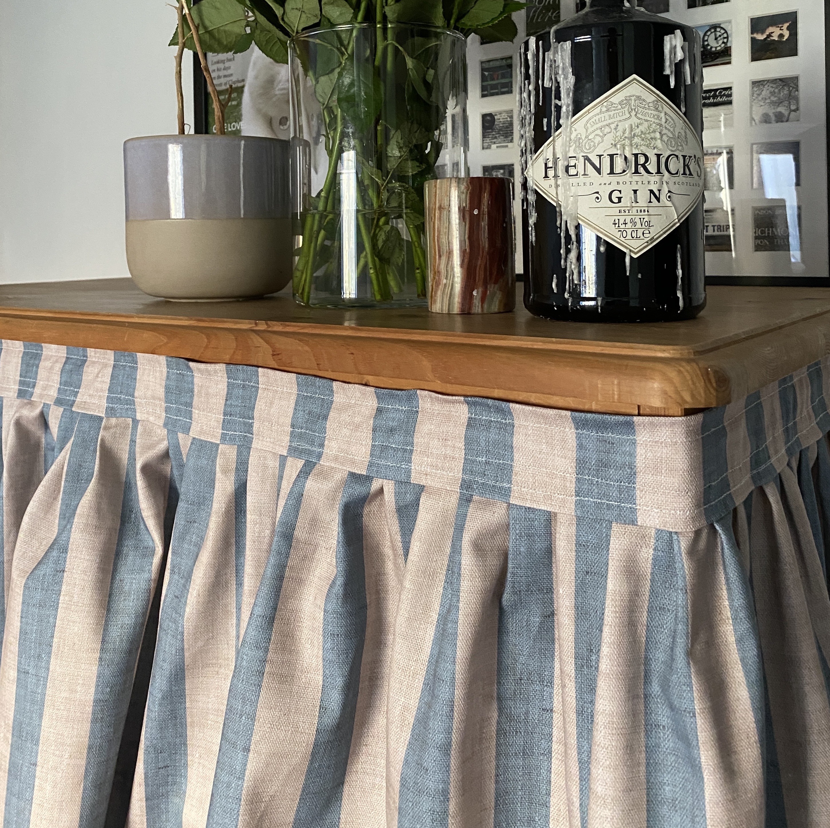 blue and beige striped pleated cabinet curtain on wooden cupboard with bottle of Hendricks gin and flowers.