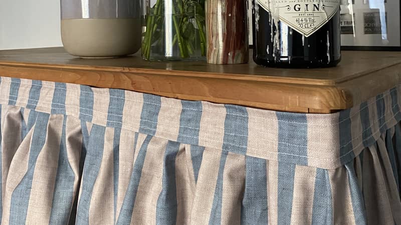 blue and beige striped pleated cabinet curtain on wooden cupboard with bottle of Hendricks gin and flowers.