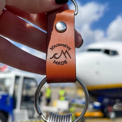 Female holding a tan Journeys Made leather travel keyring with stainless steel tokens on it, black painted nails, and a Ryanair airplane in the background