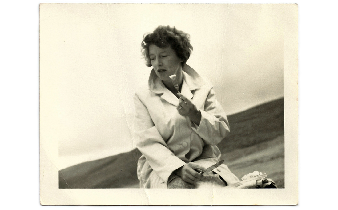 A vintage black and white photograph of a 1960s woman wearing an overcoat and eating at a picnic.