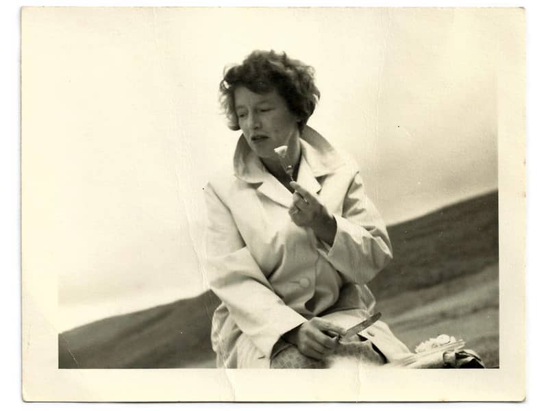 A vintage black and white photograph of a 1960s woman wearing an overcoat and eating at a picnic.