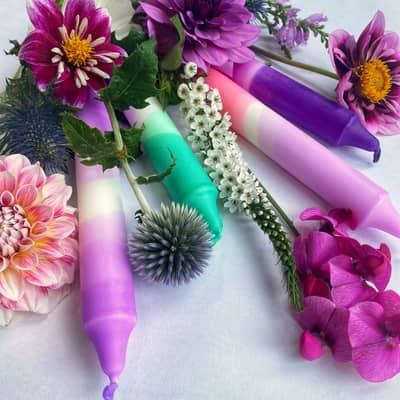 Four candles in purples, pinks and greens are positioned in a fan in amongst purple flowers.