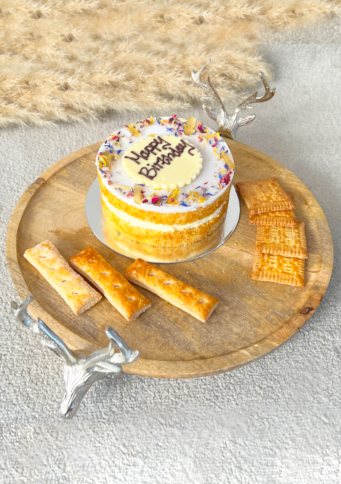 A wooden tray with stag head metal handles laid out with a birthday cake, biscuits and pastries. It is placed beautifully next to a dreamy pampas grass.