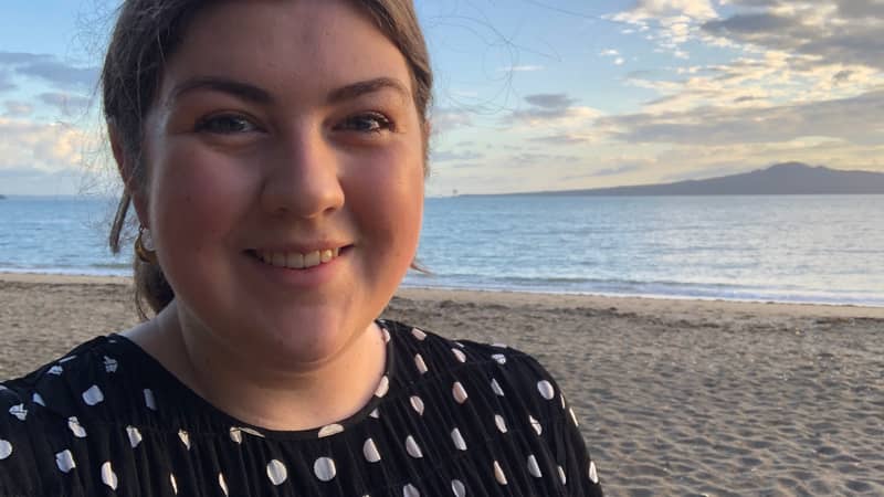 Woman standing on a beach with a cloudy blue sky in the background.
