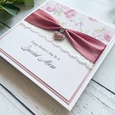 Mother's Day Greetings Card. White Square Card with Dusky Pink Border. Floral paper with decorative lace and ribbon with a heart shaped charm.