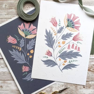 Two prints of folk art flowers in blue and pink, with a green ribbon and a pair of embroidery scissors.