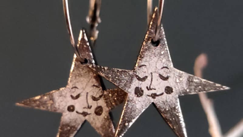 A pair of silver hoop earrings with black and grey stars with faces hanging from them. The background is dark grey and the earrings hang from white twigs.