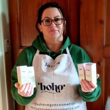 a lady with black hair tied back and red glasses wearing a green hoodie and pink apron. The apron says jackie and boho get your glow on