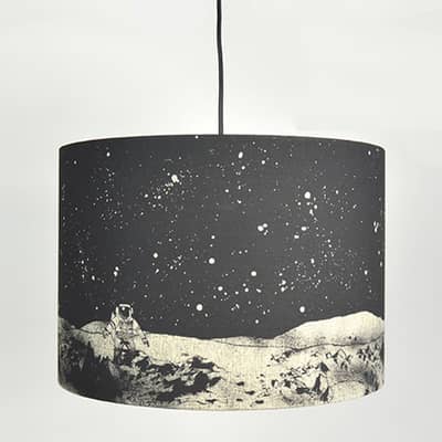 A large drum lampshade hanging on a black electrical wire. The lampshade is black and grey, with an image of an astronaut on the moon on it.