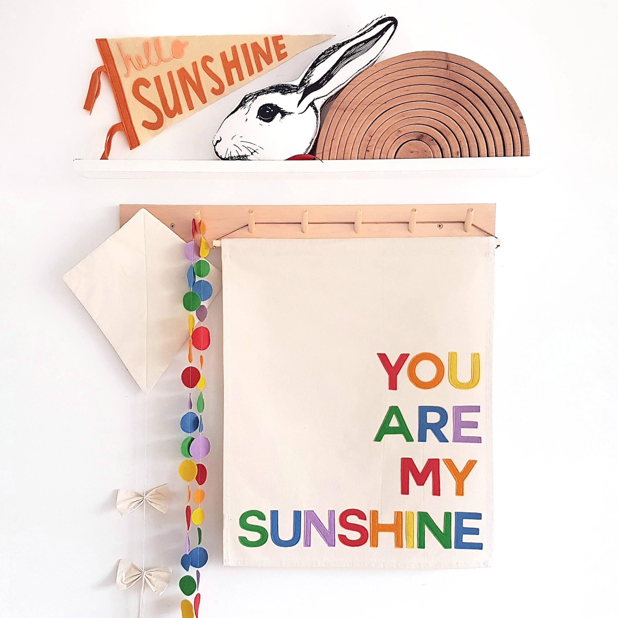 fabric banner with text " BE KIND" in rainbow colours. hanging froma peg rail with rainbows and rain drops surrounding it