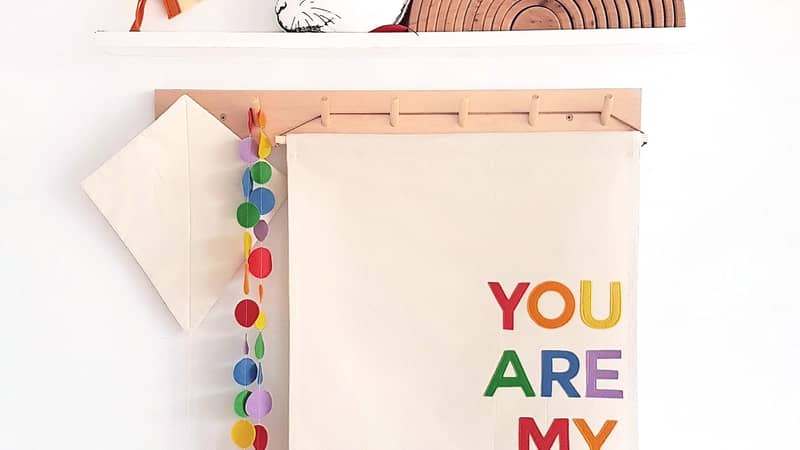 fabric banner with text " BE KIND" in rainbow colours. hanging froma peg rail with rainbows and rain drops surrounding it