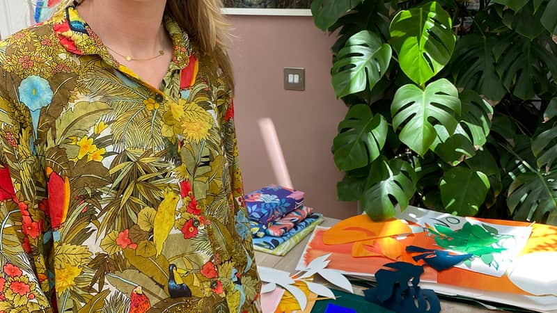 Portrait of Charlotte from Hot House Collection standing in front of her desk with art materials on. There is a big monstera plant in the background.