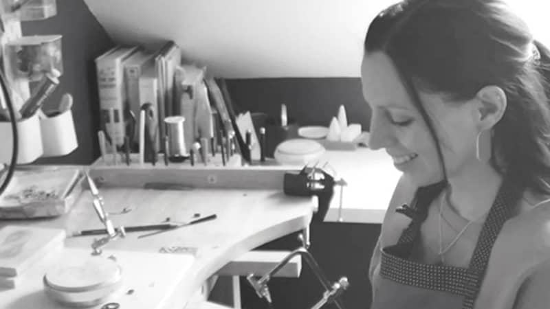 Black and white photo of jewellery designer Jodie King at a bench in a workshop. Jodie is holding a saw at the bench and is wearing an apron.
