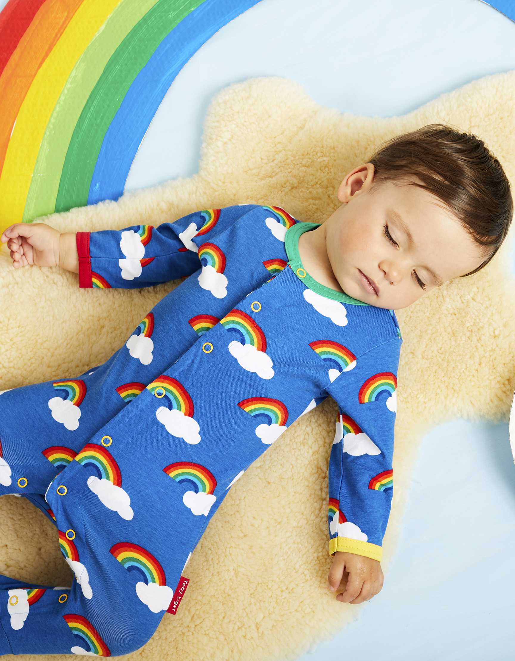 A sleeping baby wearing a Toby Tiger Organic Rainbow Print Babygrow