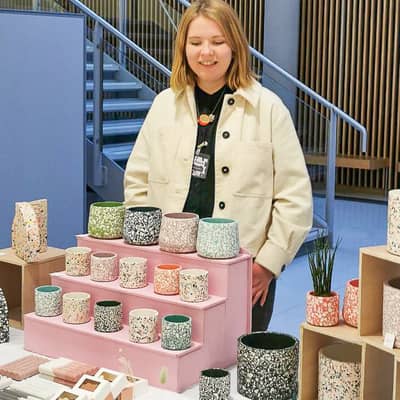 Image of Kelly, the owner and maker behind Slow Make Studio. Kelly is standing behind her market stall, which features colourful plant pots, soap dishes etc.