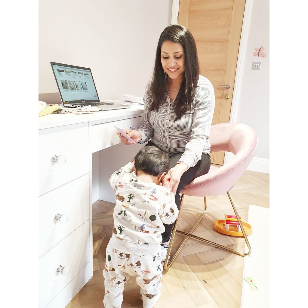 mother sitting on a chair working on her laptop with her baby standing next to her holding her hand