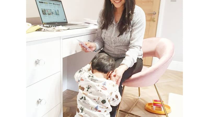 mother sitting on a chair working on her laptop with her baby standing next to her holding her hand
