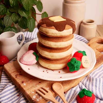 A stack of four pretend play felt food pancakes topped with syrup and butter surrounded by strawberries and banana slices.