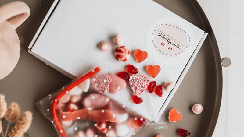 Red and white sweets pouring out of a clear cellophane bag with white spots, onto a table with our signature white gift box in the background