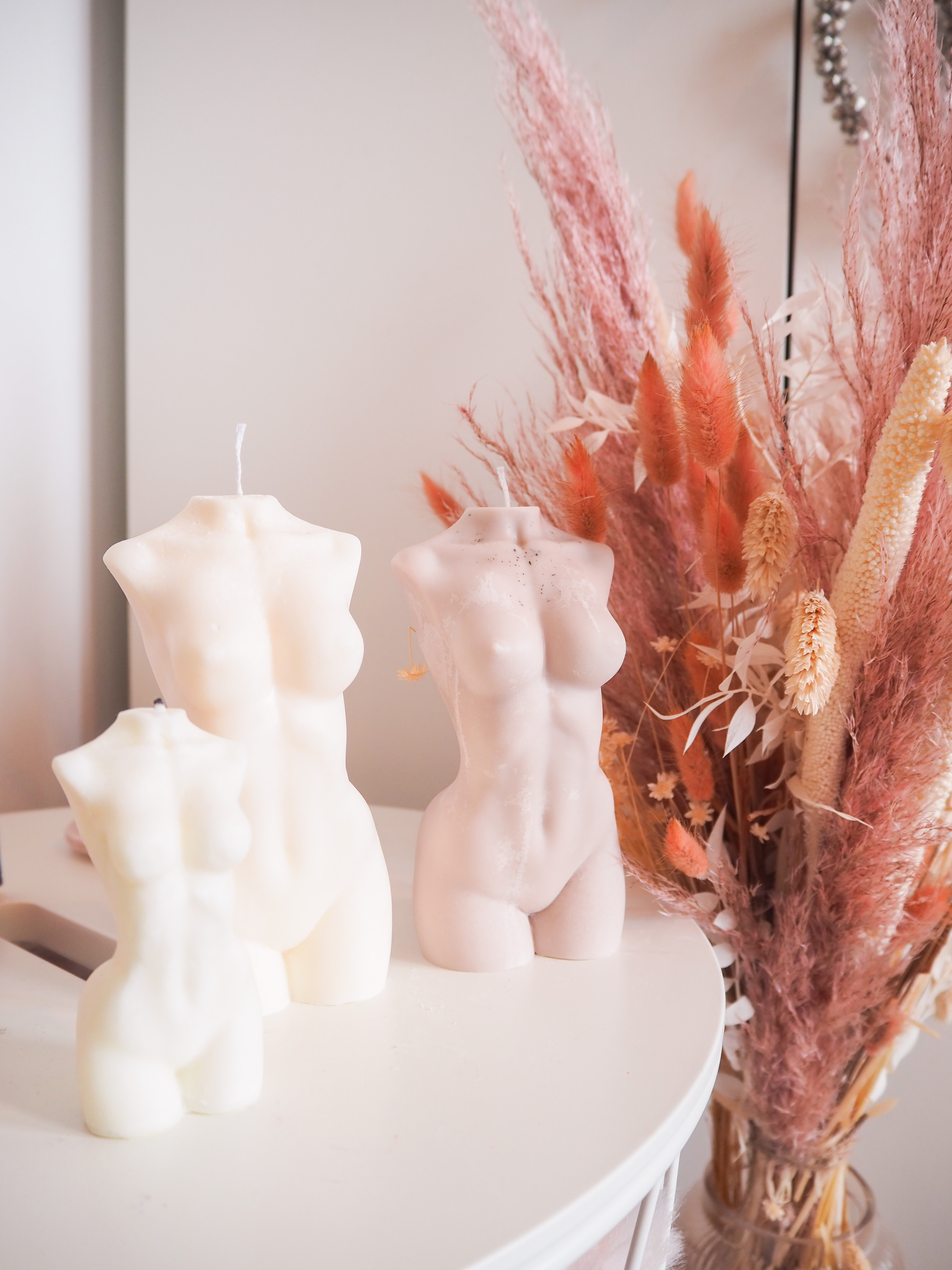 Two large Grand Grace candles next to a smaller one, stood on a table with a dried flower display in the background