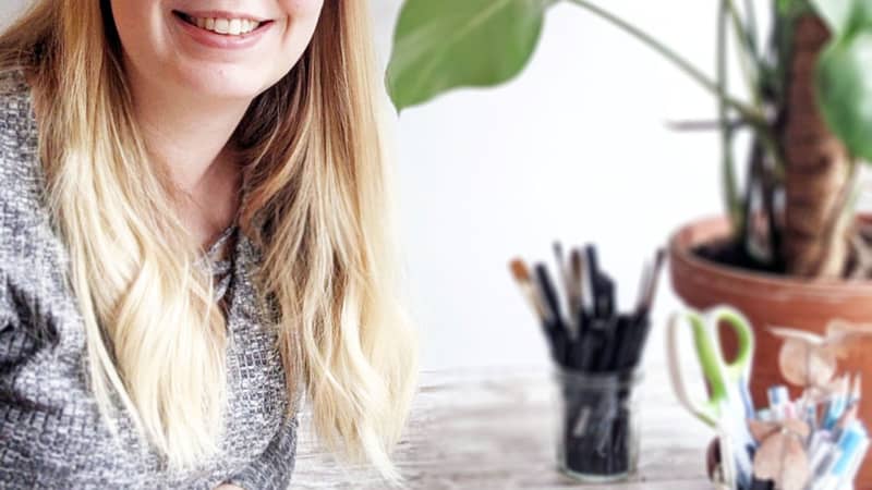 Eva sits painting at a table; behind her are a plant and two pots of pens.
