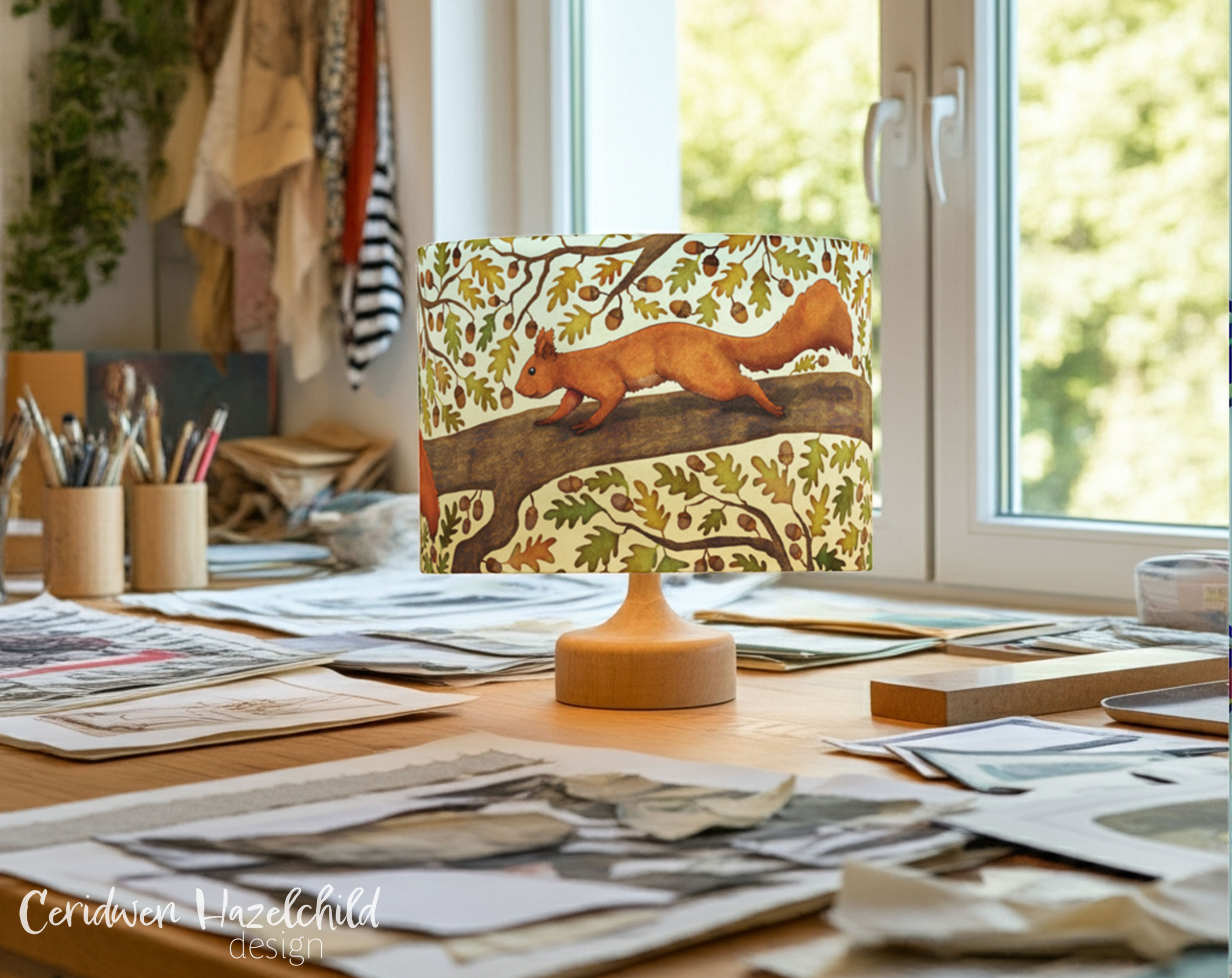 Squirrel lampshade on a table lamp base in creative studio with sketches, paper, and greenery outside window – handmade woodland decor.