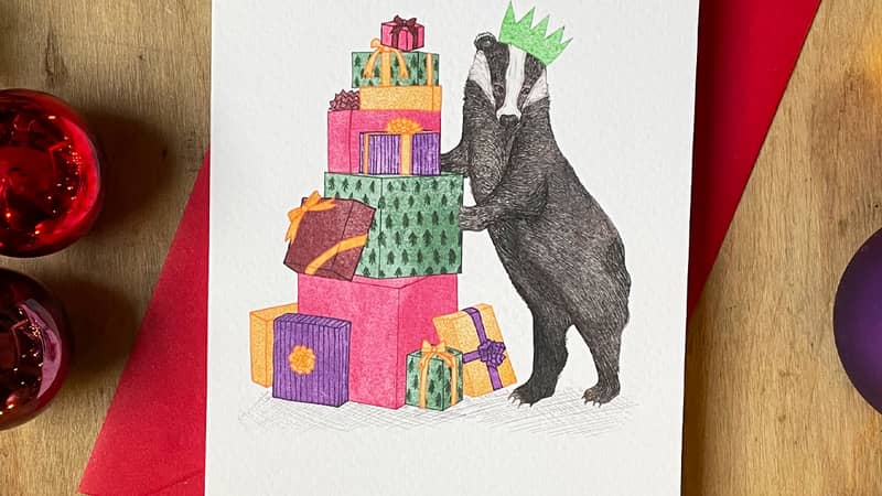A hand drawn illustration on a greeting card of a festive badger wearing a paper Christmas hat and standing besides a pile of colourful Christmas presents