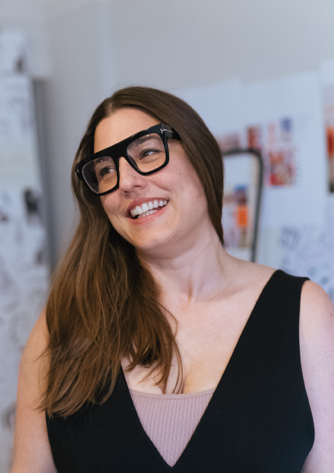 Woman with long brown hair and heavy black glasses, smiling and looking to the side.