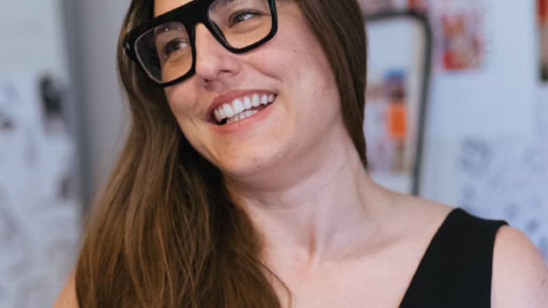 Woman with long brown hair and heavy black glasses, smiling and looking to the side.