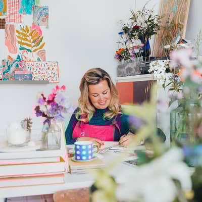 Floradore artist Lucy Innes Williams at work in her studio. Woman sat at desk surrounded by flowers in a studio.
