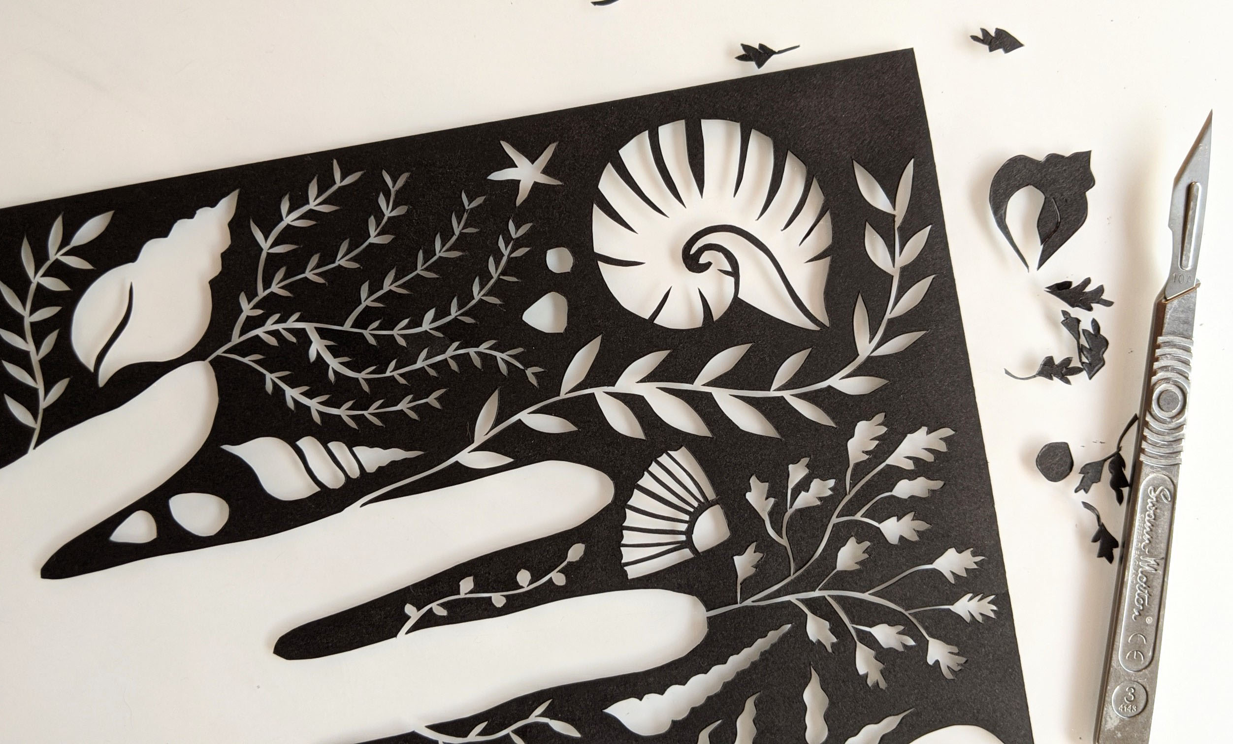 Papercut artwork of a hand surrounded by shells and seaweed, cut from black paper. Next to the artwork is a craft knife and some scraps of paper.
