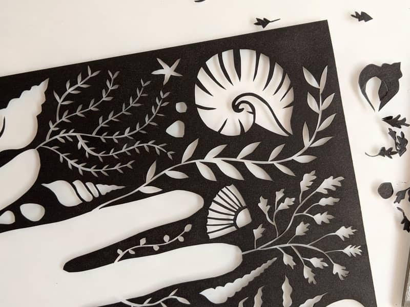 Papercut artwork of a hand surrounded by shells and seaweed, cut from black paper. Next to the artwork is a craft knife and some scraps of paper.