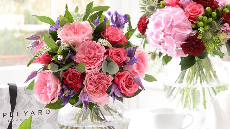 Two luxury flower bouquets in vases on a white table. The bouquets are mostly pink and include roses hydrangeas carnations and clematis