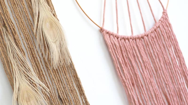 two macrame wall hangings in pink and beige with peacock feathers